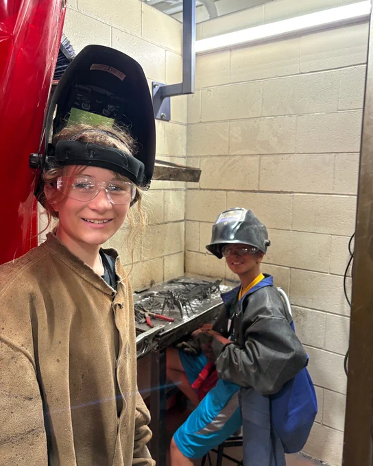 Two campers wear welding gear and smile in a Mid-State trades lab during College Camp.