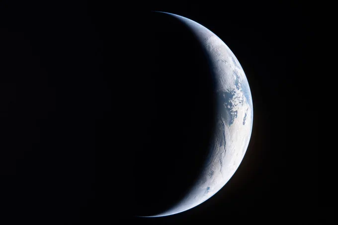 A sliver of Earth illuminated on the right side against the blackness of space, showing a thin crescent of blue oceans and white clouds while the rest of the planet is in total darkness.