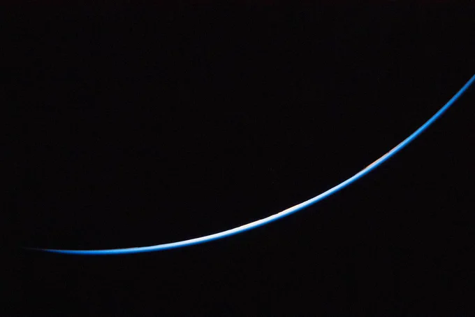 A thin, curved blue line of Earth's atmosphere brightly illuminated across the lower half of the frame against the vast darkness of space.