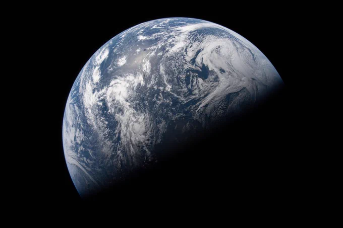 Earth illuminated against the blackness of space, with the top half showing clouds and oceans, and the bottom half fading into darkness.