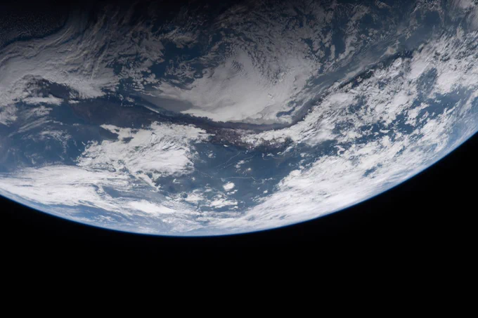 A close-up view of Earth, with swirling white clouds and blue oceans curving across the top half, and the bottom half fading into the darkness of space.