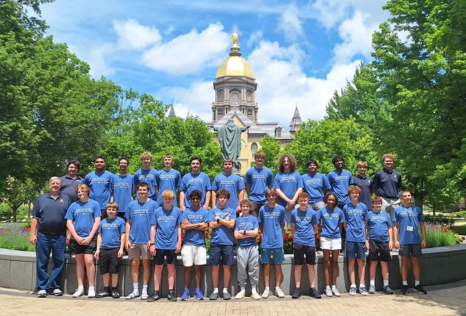 Students and instructors from the Notre Dame Engineering Innovation Hub Robotics Camp pose for a group photo in front of the University of Notre Dame’s Golden Dome, with trees and blue skies in the background.