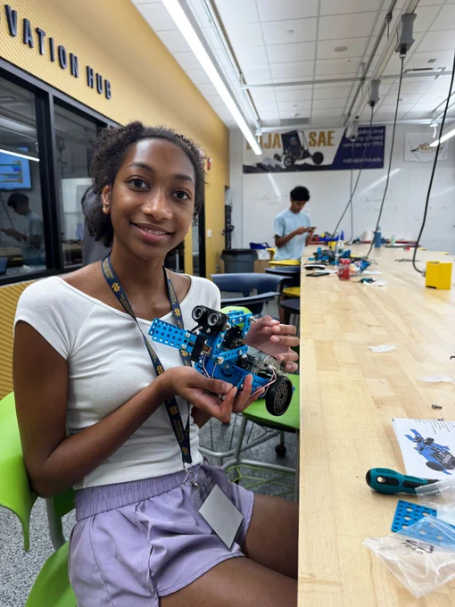 A student in the Notre Dame Engineering Innovation Hub holds a small blue robot she assembled during Robotics Camp. Parts, tools, and another student working on their robot are visible in the lab behind her.