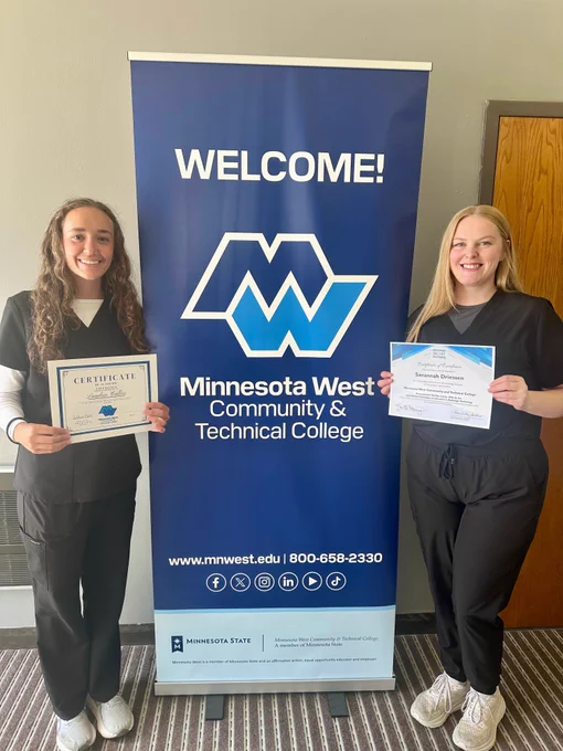 Two students in medical scrubs stand on either side of a blue Minnesota West banner that reads “WELCOME!” and displays the college logo. Each student is holding a certificate of excellence.