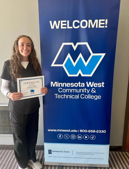 Landree stands beside a blue Minnesota West banner holding a certificate labeled “Landree Callis.” 