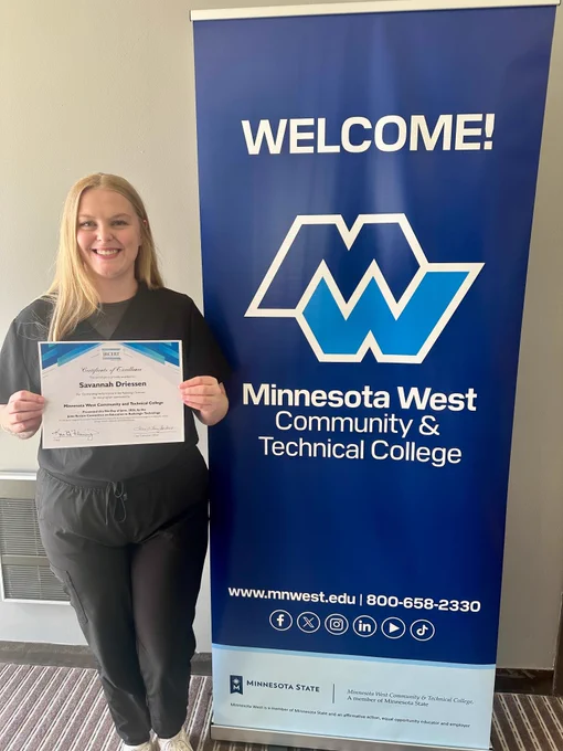 Savannah stands next to a blue Minnesota West banner and holds a certificate labeled “Savannah Driessen.” 
