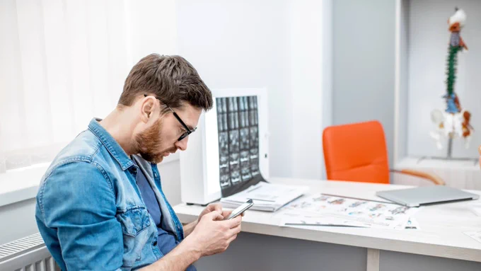 A person sits in a medical office, looking at a smartphone. There are X-ray images on a lightbox, a stack of papers on the desk, and a model of a spine in the background.