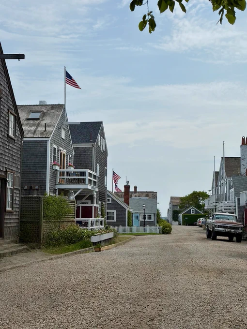 Residential gravel-road lane in Nantucket with shingled cottages, American flags, and a vintage pickup truck parked outside