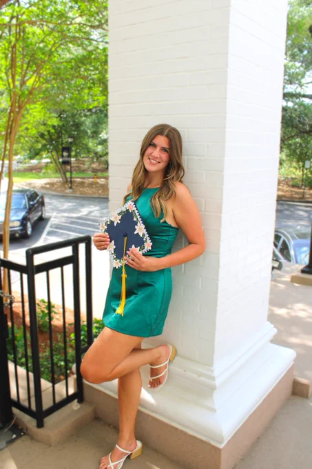A person in a green dress leans against a large white pillar, holding a decorated graduation cap with a yellow tassel. Trees and parked cars are visible in the background.
