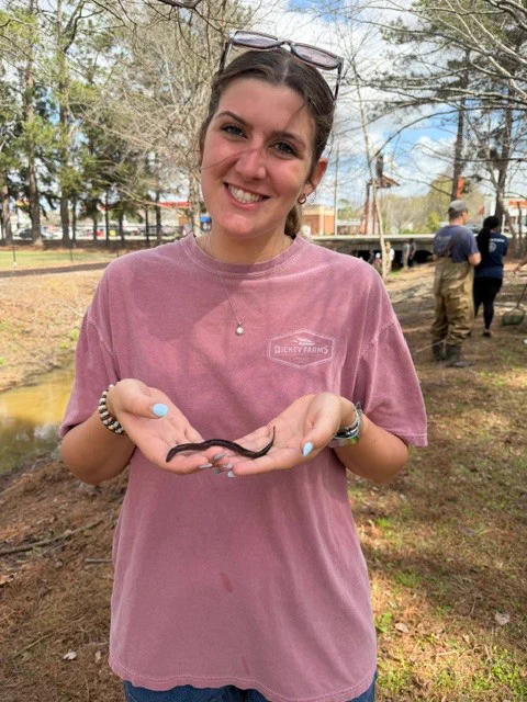 A person smiling and holding a small snake-like creature outdoors. Trees and a creek are in the background, and the person is wearing a casual pink shirt.