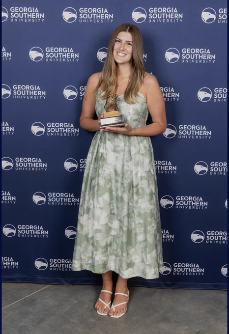 A person stands smiling while holding a trophy in front of a backdrop featuring the Georgia Southern University logo. They are wearing a light green, floral-patterned dress and sandals.