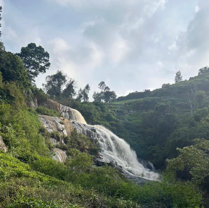 A cascading waterfall tumbles down rocky slopes, surrounded by lush greenery and distant hills under a partly cloudy sky.