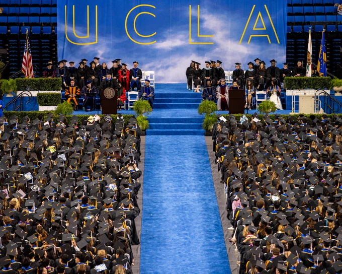 Graduation ceremony at UCLA with a large audience of graduates in caps and gowns facing a stage. The stage features faculty members seated, with a backdrop displaying a large "UCLA" sign. Flags and plants adorn the stage area.