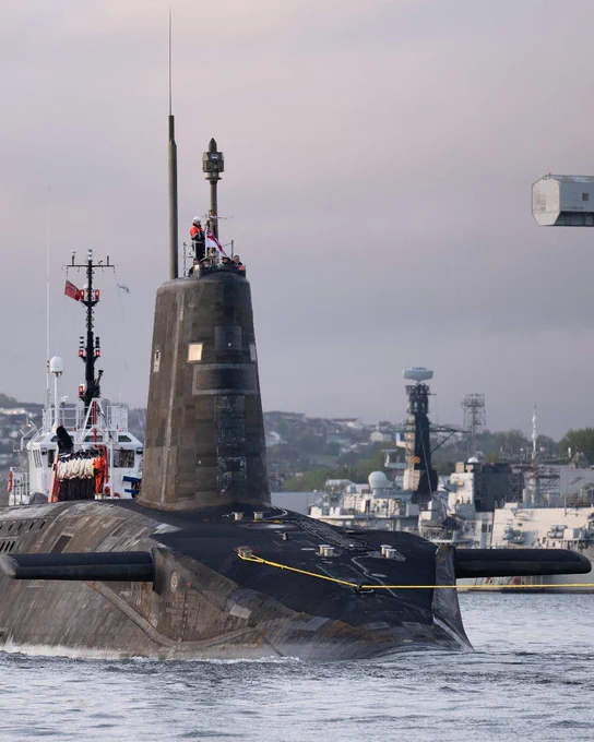 A submarine surfaced in a harbor with crew members visible on the conning tower and naval ships in the background.