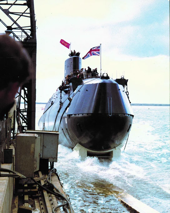 British submarine emerging from water near dock with crew on deck and Union Jack flag flying.