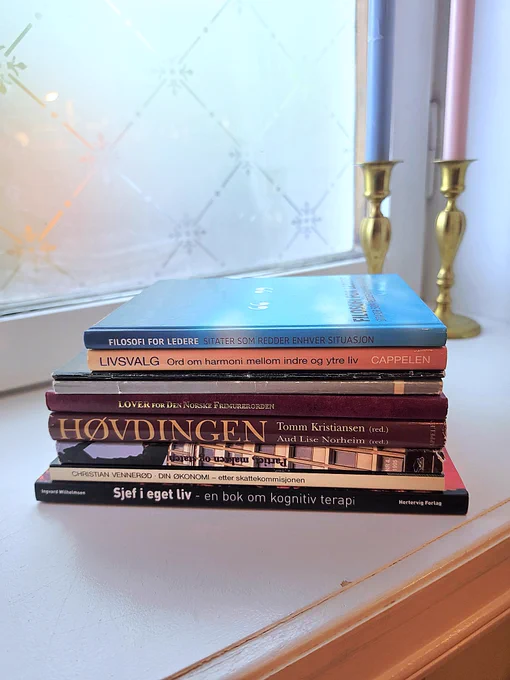 A calm stack of Norwegian books placed on a white windowsill beside two brass candle holders. Soft morning light shines through a patterned frosted window, creating a peaceful and reflective atmosphere. The books focus on philosophy, life choices, cognitive therapy, leadership, and personal growth.