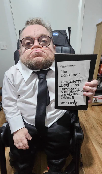 I am wearing glasses, a white shirt and black tie sits in a powered wheelchair indoors, holding a copy of the book The Department: How a Violent Government Bureaucracy Killed Hundreds and Hid the Evidence by John Pring. The book cover appears deliberately marked through, with words such as "Policy" and "Violent Government" crossed out. The man looks directly at the camera with a serious. unimpressed expression. A bookshelf and office-stvle furniture are visible in the background.