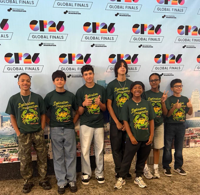 Group of seven boys wearing matching green Destination Imagination t-shirts posing in front of a CR26 Global Finals backdrop.