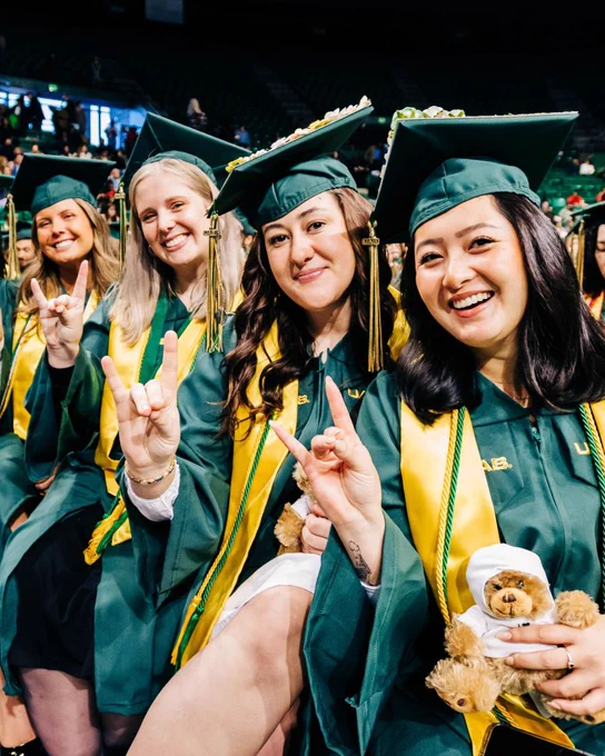 A row of smiling graduating female graduate students are giving the Blaze On hand gesture while seated in Bartow Arena.