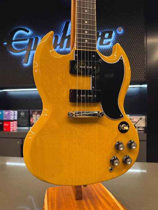 A yellow Epiphone electric guitar is displayed on a stand. The background shows a blurred Epiphone logo on the wall. The guitar features a black pickguard and several control knobs.
