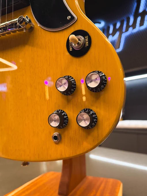 Close-up of a yellow Epiphone electric guitar body on a stand. It has four black knobs and a switch. The background features a blurry Gibson logo.