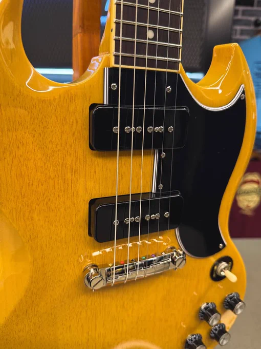A close-up of an electric Epiphone SG Special guitar, featuring a striking yellow body with black pickups and a black pickguard. The instrument has chrome hardware, including the bridge and tuning pegs. Several control knobs are visible on the lower right of the body.