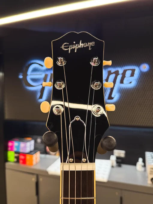 Close-up of an Epiphone guitar headstock in a store. The background displays the illuminated Epiphone logo, with blurred shelves containing boxes and products.
