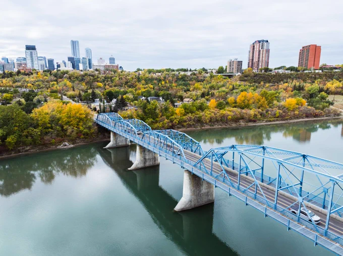 A blue steel truss bridge spans a calm river with autumn trees and a city skyline in the background under a cloudy sky.