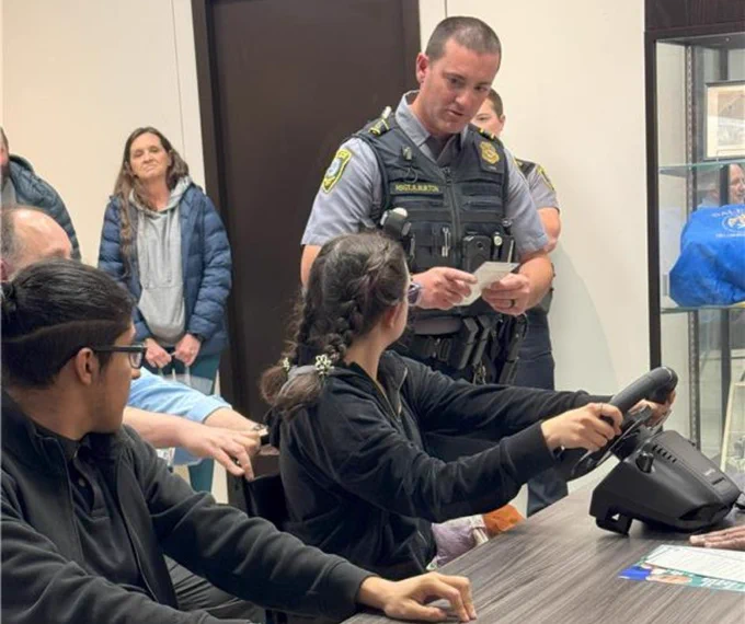 Young girl holding a steering wheel controller at a table while a police officer stands nearby with papers in hand inside a room with other people.