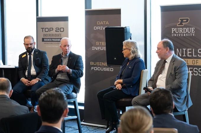 A panel of four people seated in front of banners from Purdue University's Daniels School of Business. One person is speaking into a microphone, while the others listen attentively.