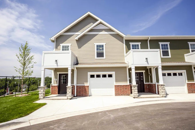 The image shows a modern townhouse complex with two-story units featuring garages, brick and siding exteriors, and small balconies. The area is neatly landscaped and the sky is clear.