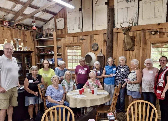 A group of people, mostly older adults, are gathered in a rustic room with wooden walls and shelves with various items, including a deer mount. Some are standing, and others are seated around a table with a tablecloth and red cups. The group is smiling and appears to be enjoying a social gathering.