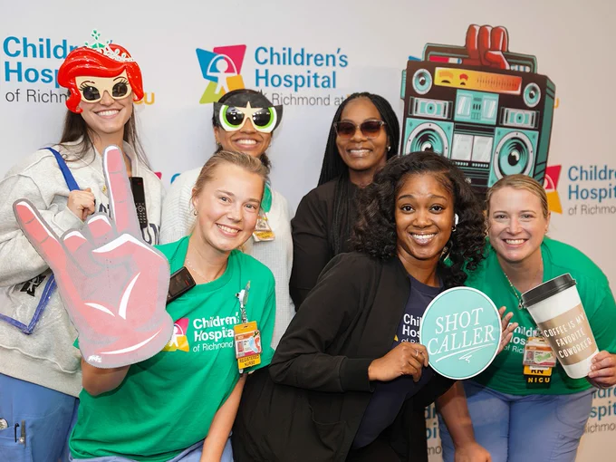 A group is posing at the Children's Hospital of Richmond event. They are smiling and holding playful props, such as a coffee cup, sunglasses, and a toy boombox. The backdrop features the hospital's logo.