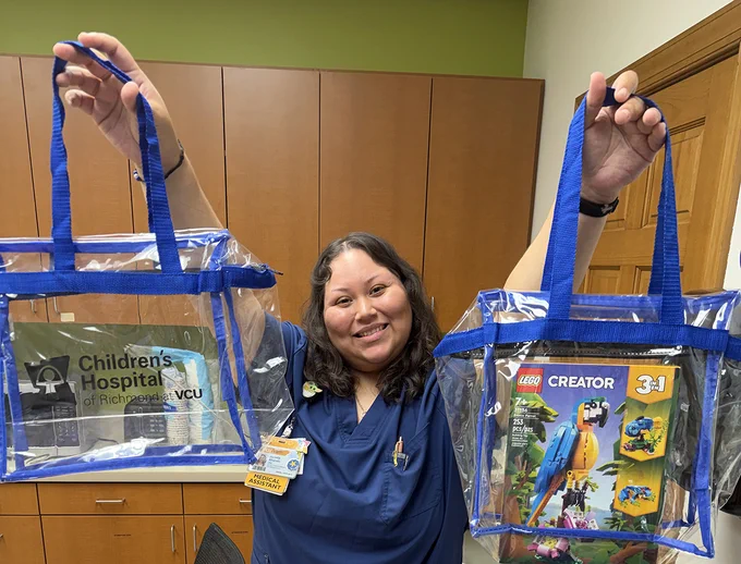 A person smiles while holding two clear blue-handled bags. One bag has a Children's Hospital of Richmond at VCU logo, and the other contains a LEGO Creator set. The person is standing in a room with wooden cabinets and a green wall.