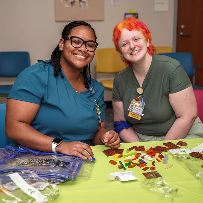 Two individuals smile for the camera while sitting at a table with LEGO bricks.
