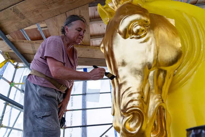 A craftsperson stands on scaffolding applying gold leaf to the head of a large gilded horse sculpture during restoration work.
