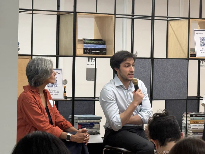 Two people are sitting and speaking in a library setting, with bookshelves in the background. One person is holding a microphone.