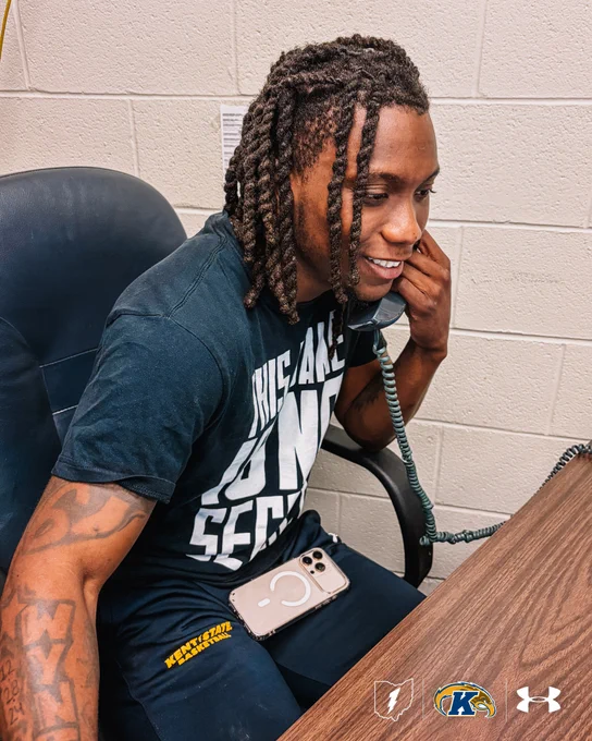 Kent State Men's Basketball player Jamal Sumlin sits at a desk, smiling while speaking on a corded office telephone. He wears a dark t-shirt reading "This Game Is No Secret" and navy Kent State Basketball sweatpants. The Kent State, Ohio, and Under Armour logos appear in the bottom right corner.