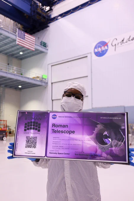 A photo of an individual in a clean room at NASA's Goddard Space Flight Center, wearing full clean room protective attire. They are holding a large, purple boarding pass that reads "Roman Telescope." Credit: NASA