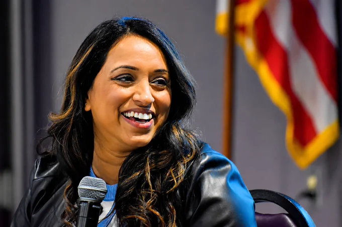 A woman with long dark hair smiles while seated indoors; an out-of-focus American flag is visible in the background.