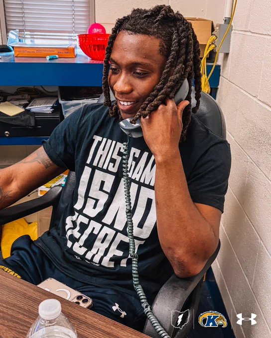 Kent State Men's Basketball player Jamal Sumlin smiles while talking on a corded office telephone, seated at a desk with a water bottle and his smartphone nearby. He wears a dark "This Game Is No Secret" t-shirt and Under Armour shorts. The Kent State, Ohio, and Under Armour logos appear in the bottom right corner.