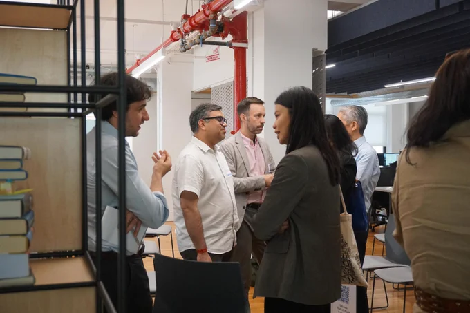A group of people are engaged in conversation in an office space. Books are visible on a shelf in the foreground. A red pipe runs along the ceiling.