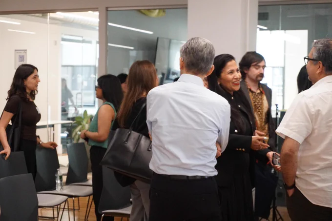 A group of people are engaged in conversation at an indoor event or meeting. They are standing and some are holding bags. There are chairs and tables in the background, with a large window and glass wall separating different areas of the room.