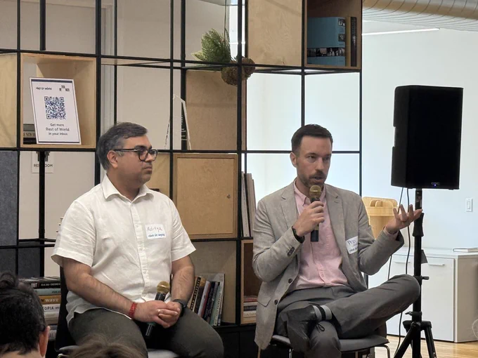 Two individuals are seated and speaking into microphones during an event. They are in front of a metal shelf with a QR code on display. The setting is a well-lit, modern office space.