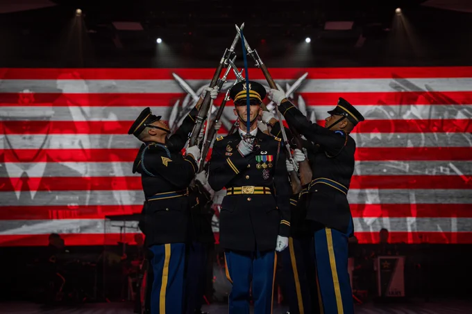 The U.S. Army Drill Team performs a precision routine in dark blue dress uniforms during the U.S. Army\u2019s 251st Birthday Twilight Tattoo at Joint Base Myer-Henderson Hall, Virginia, June 10, 2026. Four Soldiers cross their bayonet-tipped rifles to form an arch over a central Soldier who is standing sharply at attention, holding a sword vertically in front of his face. A vibrant American flag is projected on the screen behind them.