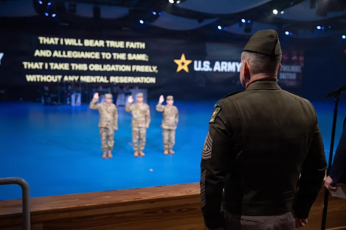 Viewed from behind, a senior U.S. Army enlisted leader in an Army Green Service Uniform observes three Soldiers on a blue stage who are raising their right hands to take an oath during the U.S. Army\u2019s 251st Birthday Twilight Tattoo at Joint Base Myer-Henderson Hall, Virginia, June 10, 2026. A massive screen behind the Soldiers projects the text of the Oath of Enlistment alongside the U.S. Army logo.