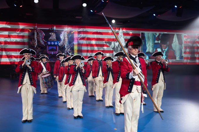 The U.S. Army Old Guard Fife and Drum Corps marches in formation across a blue floor during the U.S. Army\u2019s 251st Birthday Twilight Tattoo at Joint Base Myer-Henderson Hall, Virginia, June 10, 2026. They are wearing historical red coats, white trousers, and black tricorne hats. A Drum Major leads the group holding a tall spontoon, followed by musicians playing fifes and drums. A large American flag graphic is displayed in the background.