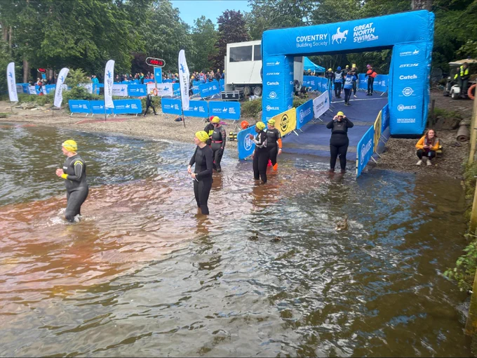 Participants in wetsuits and swim caps exit a lake at an event marked by banners and signs for the Coventry Building Society and Great North Swim. Spectators are visible in the background.