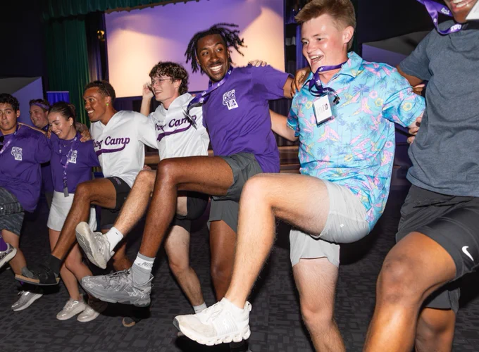 A group of people wearing t-shirts that say "Frog Camp" and colorful attire are lined up with arms around each other, kicking their legs in unison. They are smiling and appear to be enjoying an energetic activity indoors.