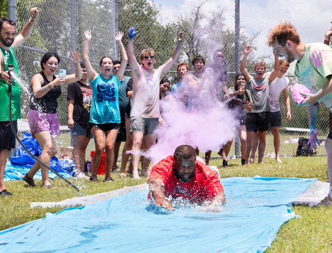 A person in a red shirt slides on a blue tarp covered in water and splashes through purple powder, surrounded by an enthusiastic group of people cheering. The setting is outdoors on grass with a chain-link fence and trees in the background.